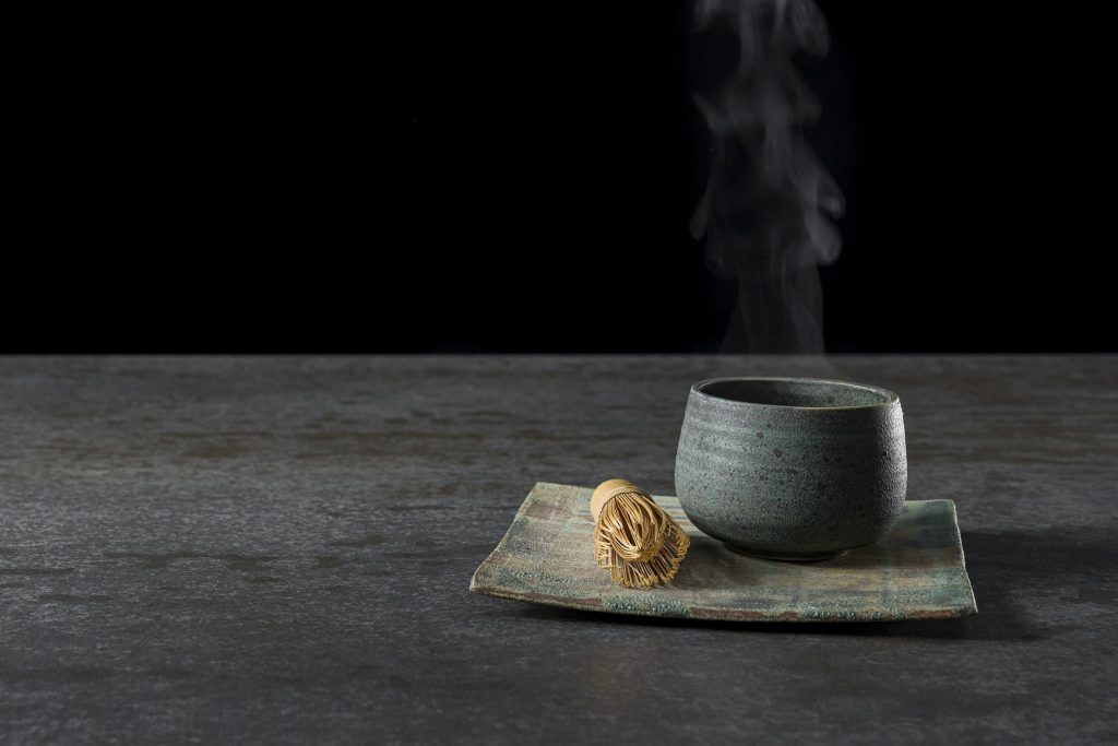 Koyu renkli, bakır tonlu Ascale Millenium Copper porselen tezgah üzerinde buharı tüten çay fincanı ve bambu çırpıcı.
