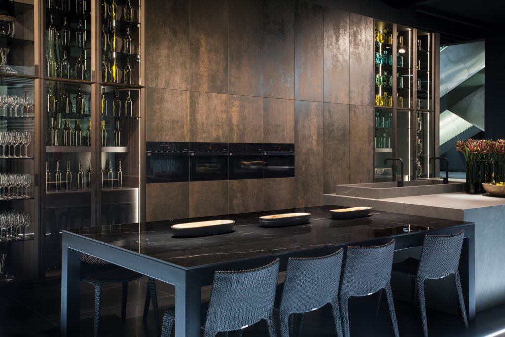 Paslanmış metal görünümlü Ascale Moma Rusteel porselen panellerle kaplı modern mutfak dolapları ve siyah yemek masası.